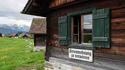 Traditionelles Chiemgauer Holzhaus mit grün gestrichenen Fensterläden und einem Schild Ferienwohnung zu vermieten vor Alpengipfeln